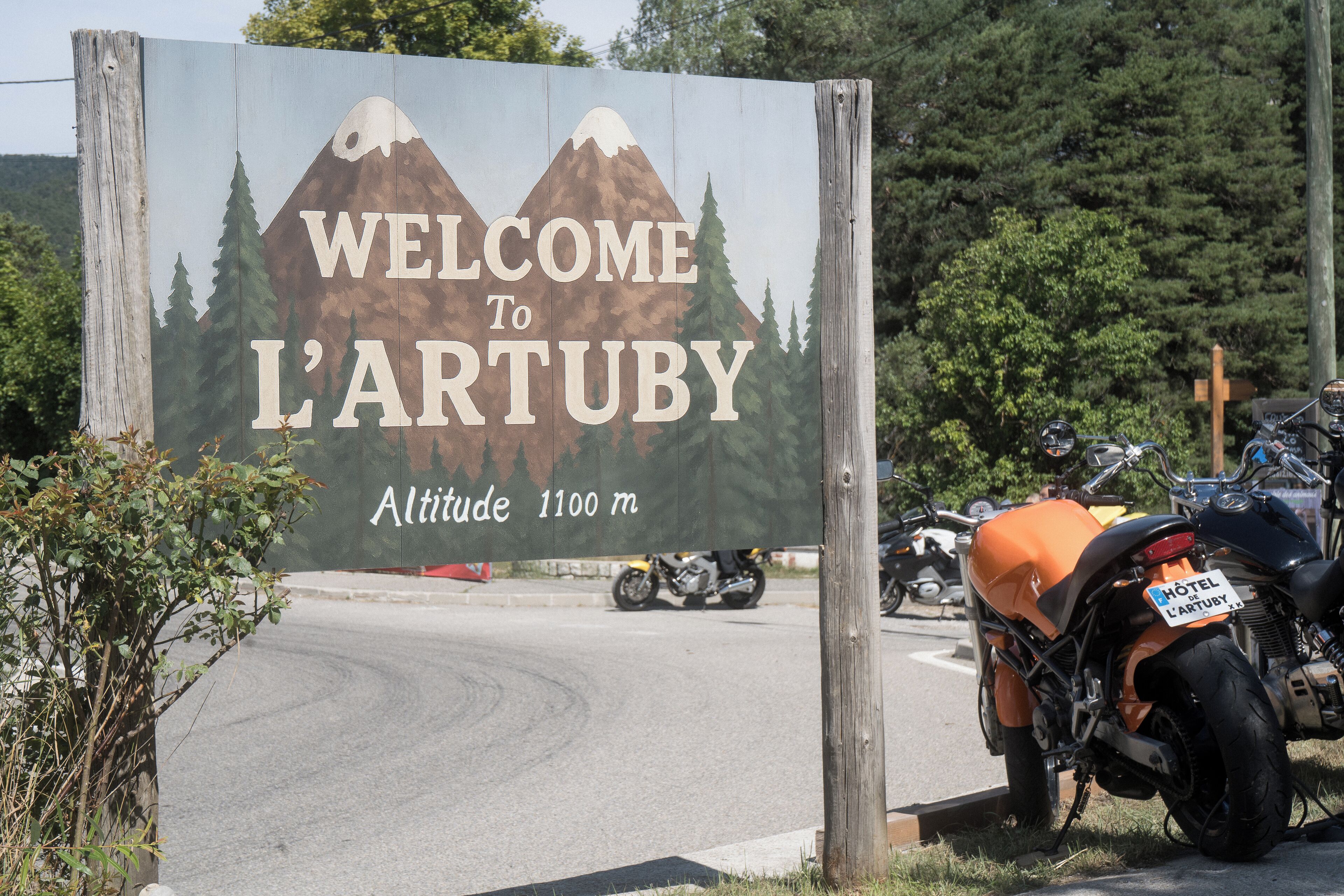 Señal de bienvenida de L'Artuby con un paisaje de montaña y bosque, y una motocicleta estacionada cerca.