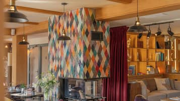 A modern interior with a geometric-patterned wall, a fireplace, a dining area with a glass table, and a cozy seating area with a sofa and bookshelves.