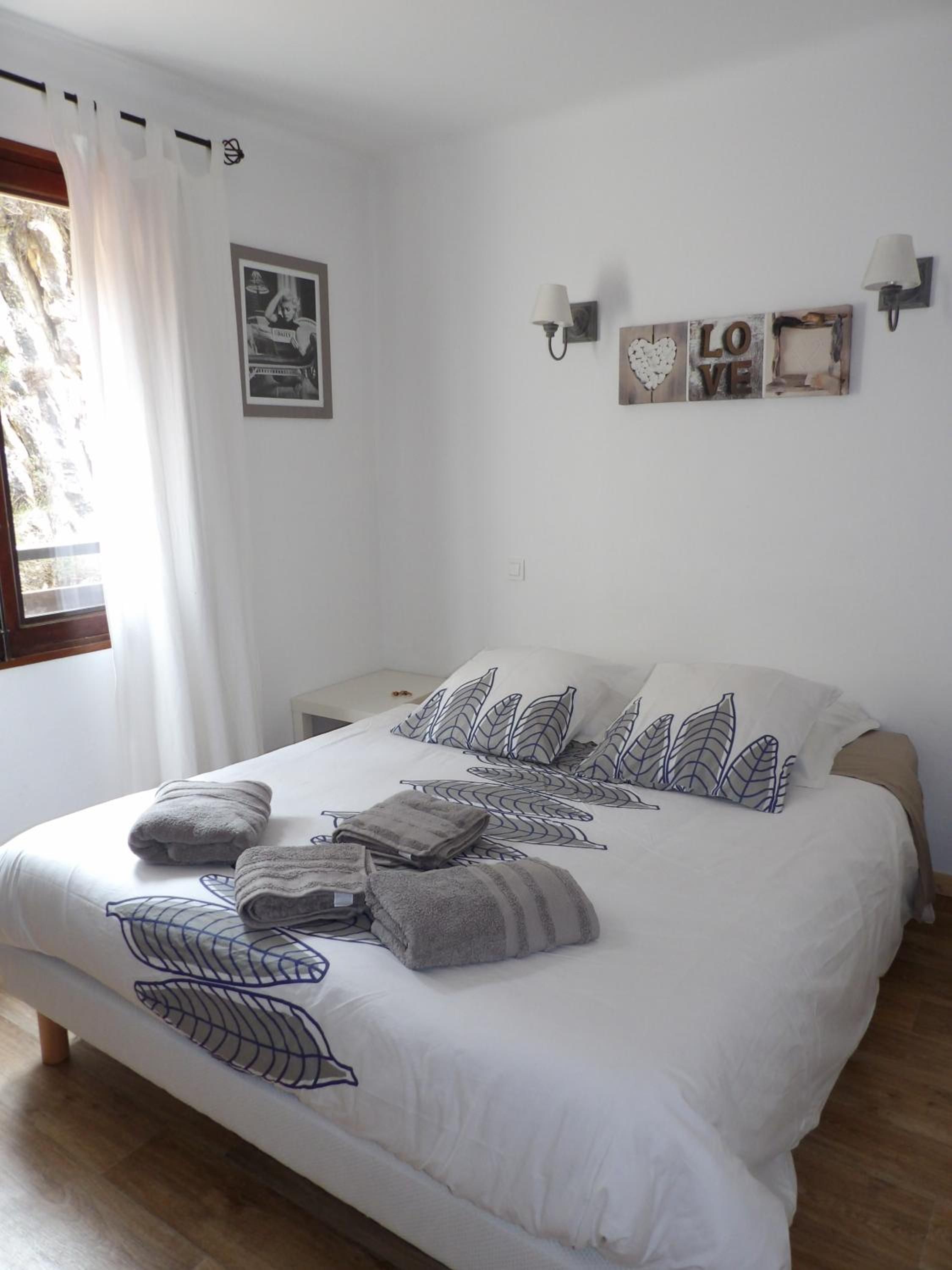 Una cama bien hecha con almohadas y toallas, una pared con un cartel que dice 'LOVE', y una ventana con cortinas.