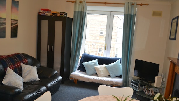 A living room with a black leather sofa, a white armchair, a small round table with a plant, and a television on a stand. - Abbey Street City Centre Apartment (Dublin)
