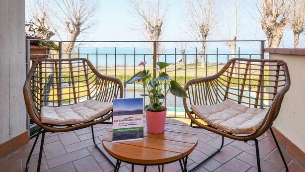 Two wicker chairs with cushions on a balcony with a view of the sea and trees. - My Sirmione Beach Apartment Superior (Sirmione)