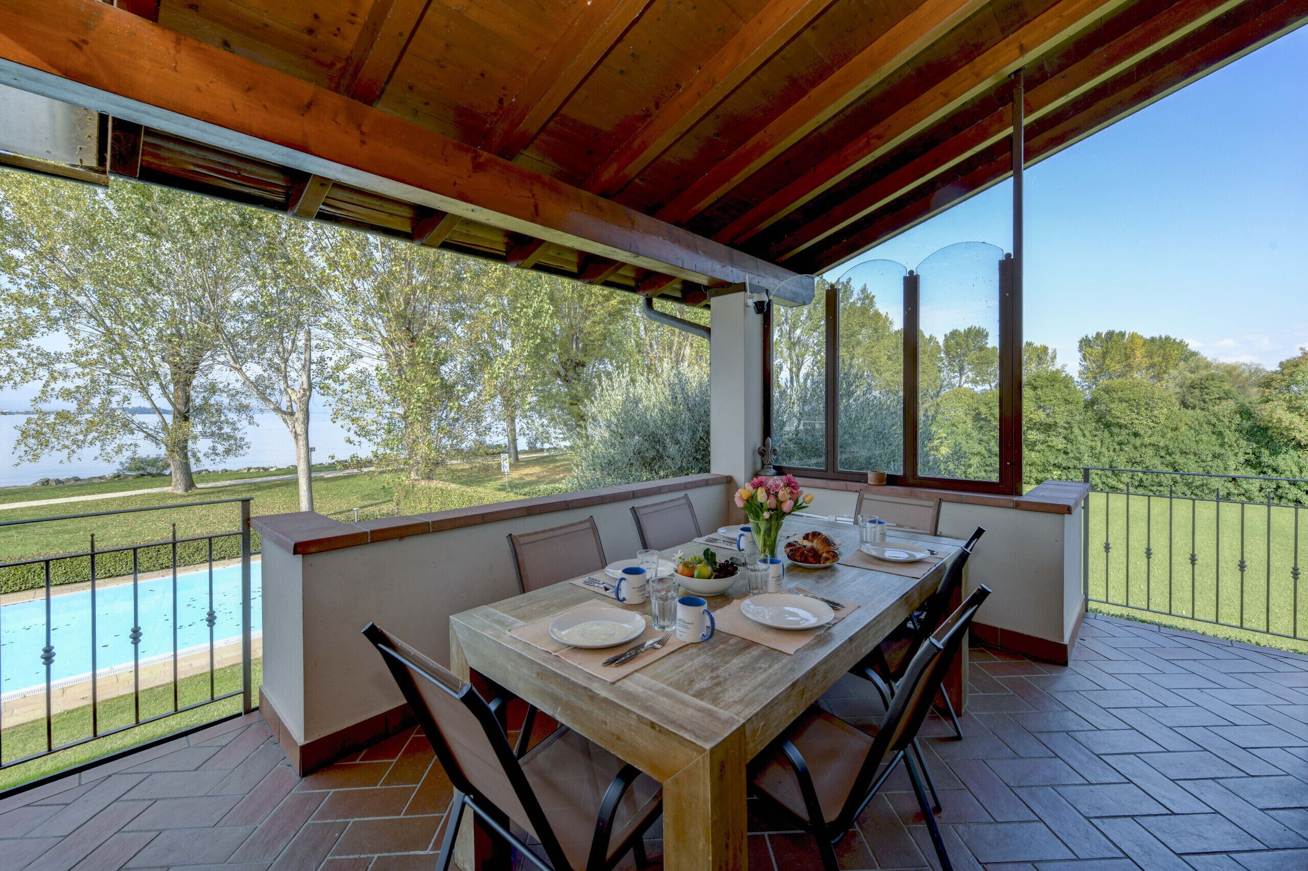 An outdoor dining area with a wooden table set for four, overlooking a swimming pool and lush greenery.