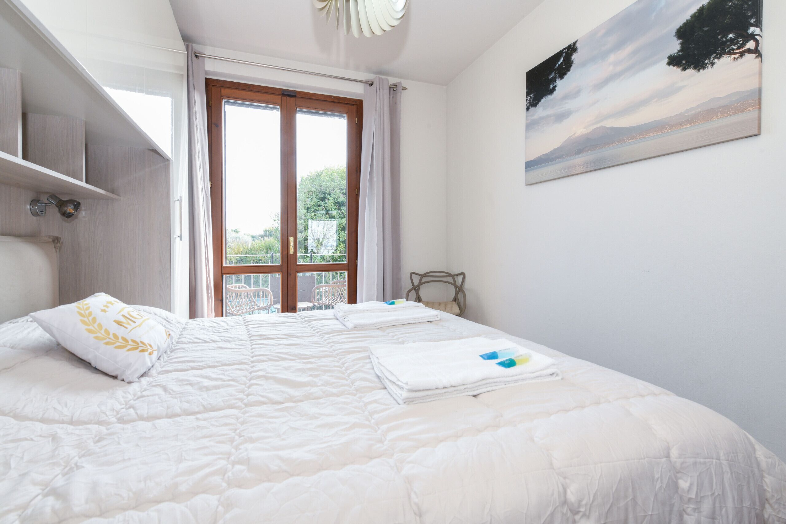 A bedroom with a large bed, a view of the outdoors through a window, and a painting on the wall.