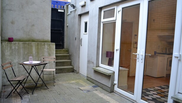 Un petit espace extérieur aménagé en patio, avec une table et des chaises, une porte donnant sur la cuisine et un mur en béton.