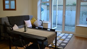 Un coin repas avec une table dressée pour deux personnes, un canapé avec des coussins, une petite table d’appoint et un tapis à motifs.