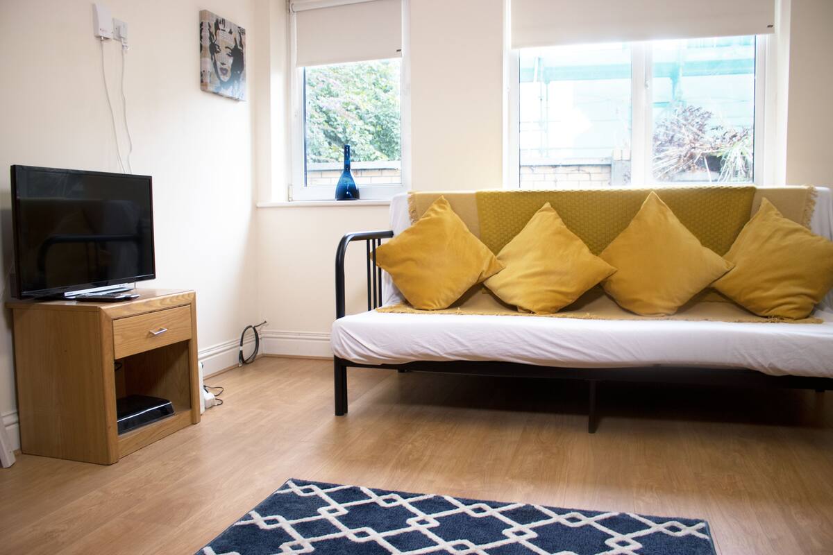 A bedroom with a bed, a TV on a wooden cabinet, and a window with a view of trees.