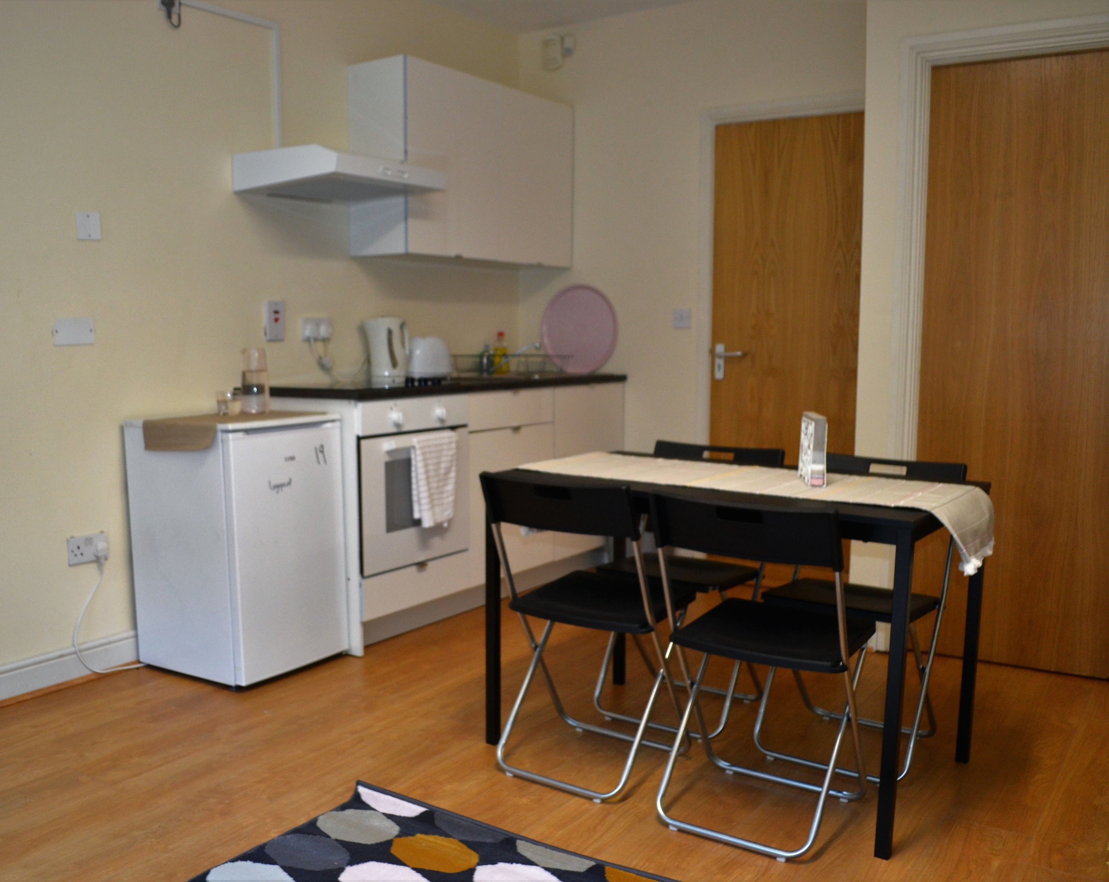 A compact kitchen with a white refrigerator, a small oven, and a coffee maker. There is a dining table with four chairs and a small rug on the floor.