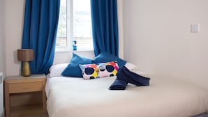 Un lit bien fait, avec une literie bleue et blanche, un coussin coloré et une serviette enroulée sur une table de chevet en bois, sur laquelle se trouve une lampe.