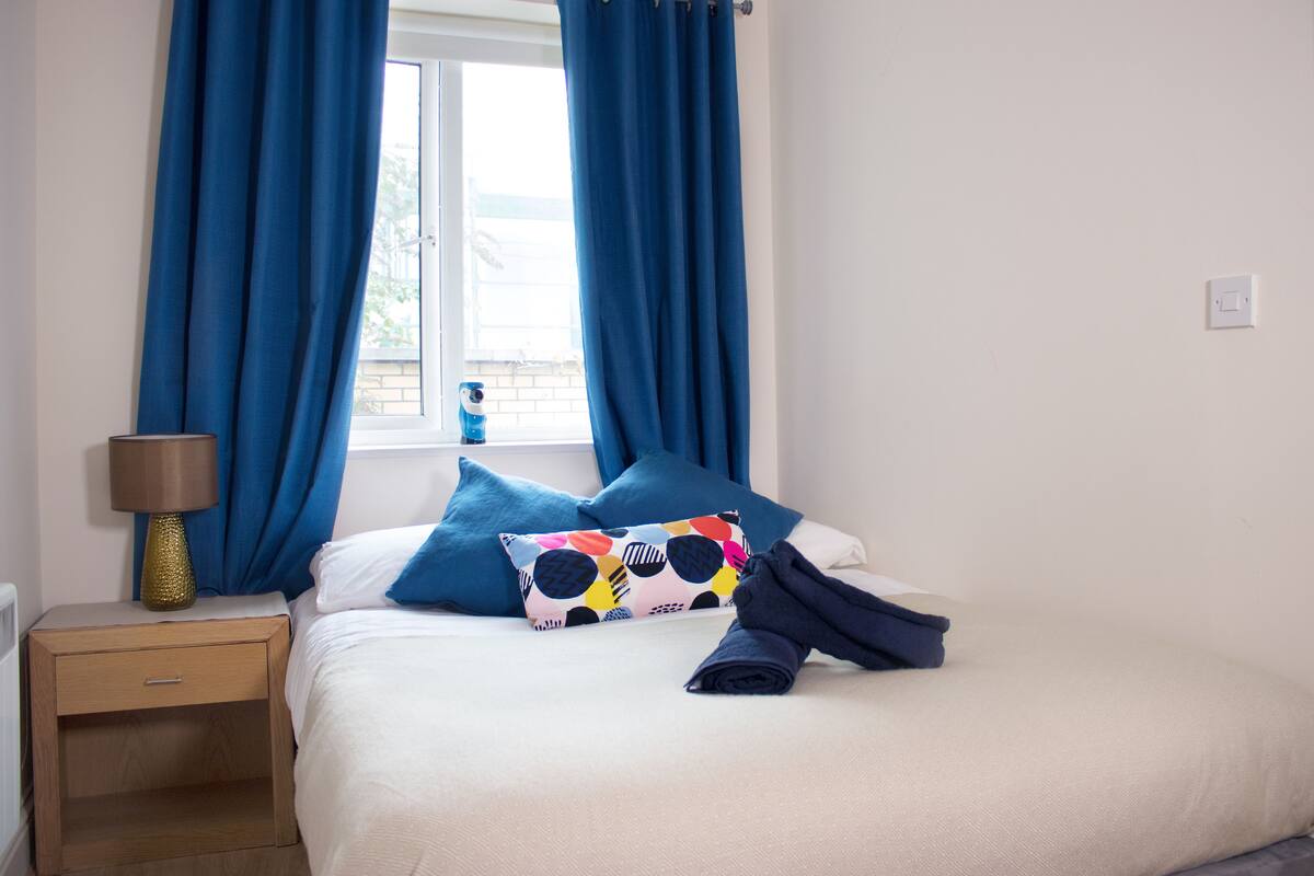A neatly made bed with blue and white bedding, a colorful pillow, and a rolled towel on a wooden nightstand with a lamp.