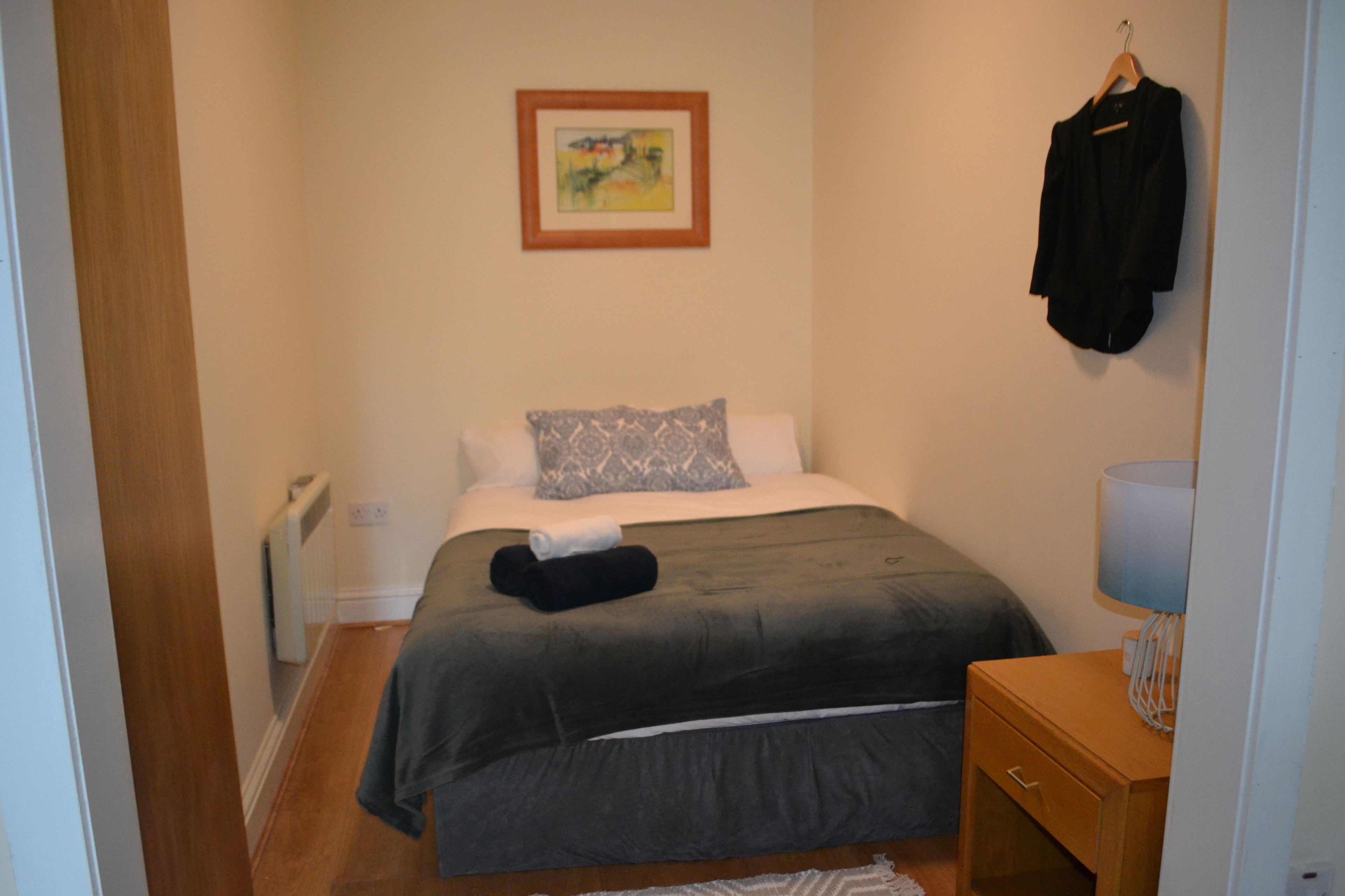 A small, neatly arranged bedroom with a bed, a bedside table, a hanging coat, and a framed picture on the wall.