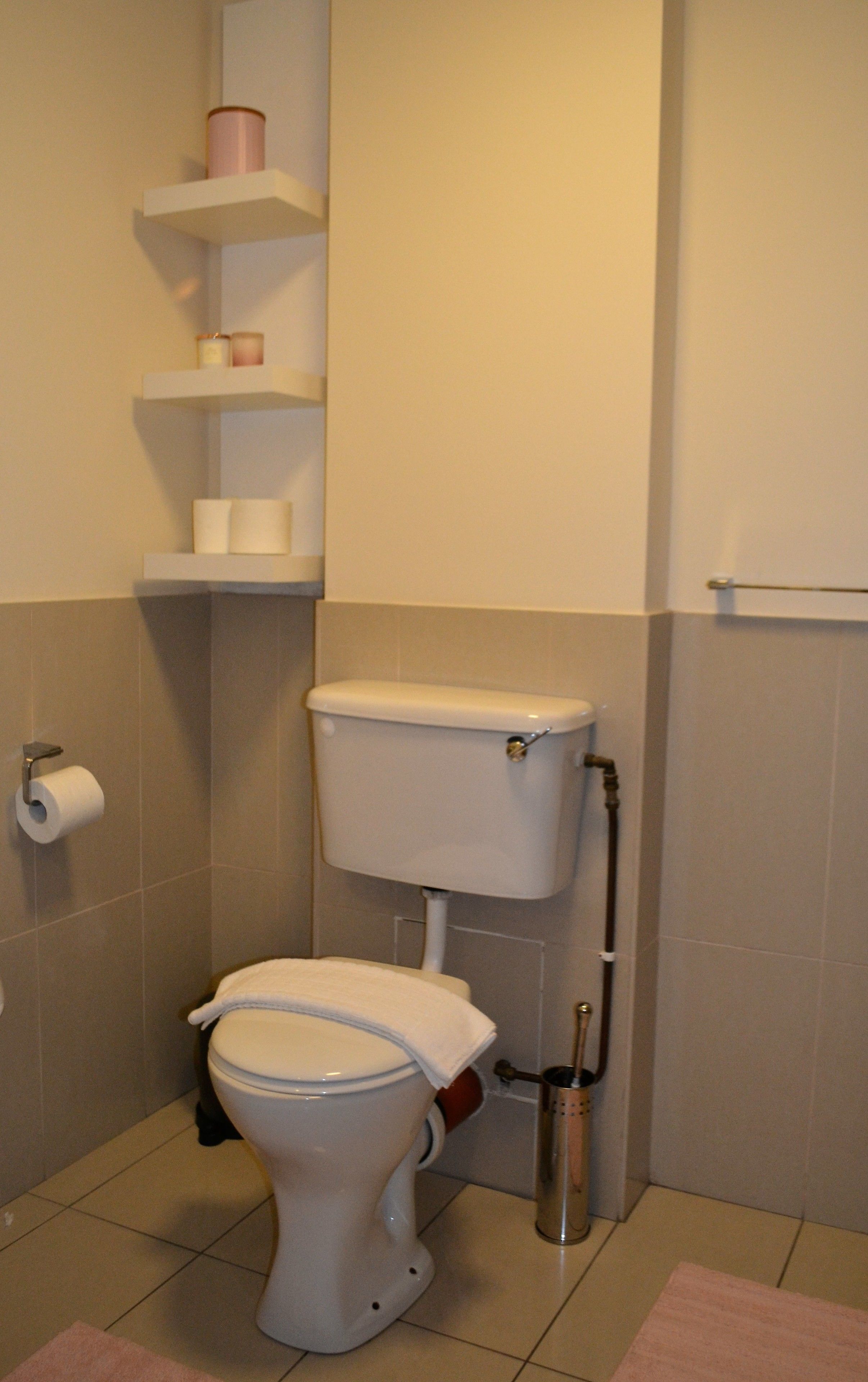 Un baño estándar con inodoro, un rollo de papel higiénico, un estante con un rollo de toallas de papel y un escobillón.