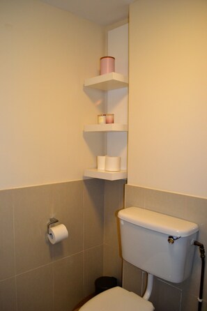 A bathroom with a toilet, a roll of toilet paper, and a shelf with a pink container and a roll of paper towels. - Cozy Modern Bedroom Apartment City L1 (Dublin)