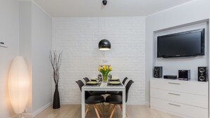 Un espace de salle à manger moderne avec un mur en briques blanches, une table à manger avec des chaises, un luminaire suspendu, un vase avec des fleurs et un téléviseur à écran plat fixé au mur.