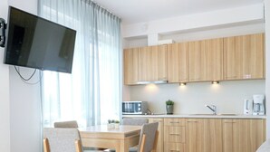 A modern kitchen with wooden cabinets, a dining table with four chairs, a flat-screen TV mounted on the wall, and a coffee maker on the counter.