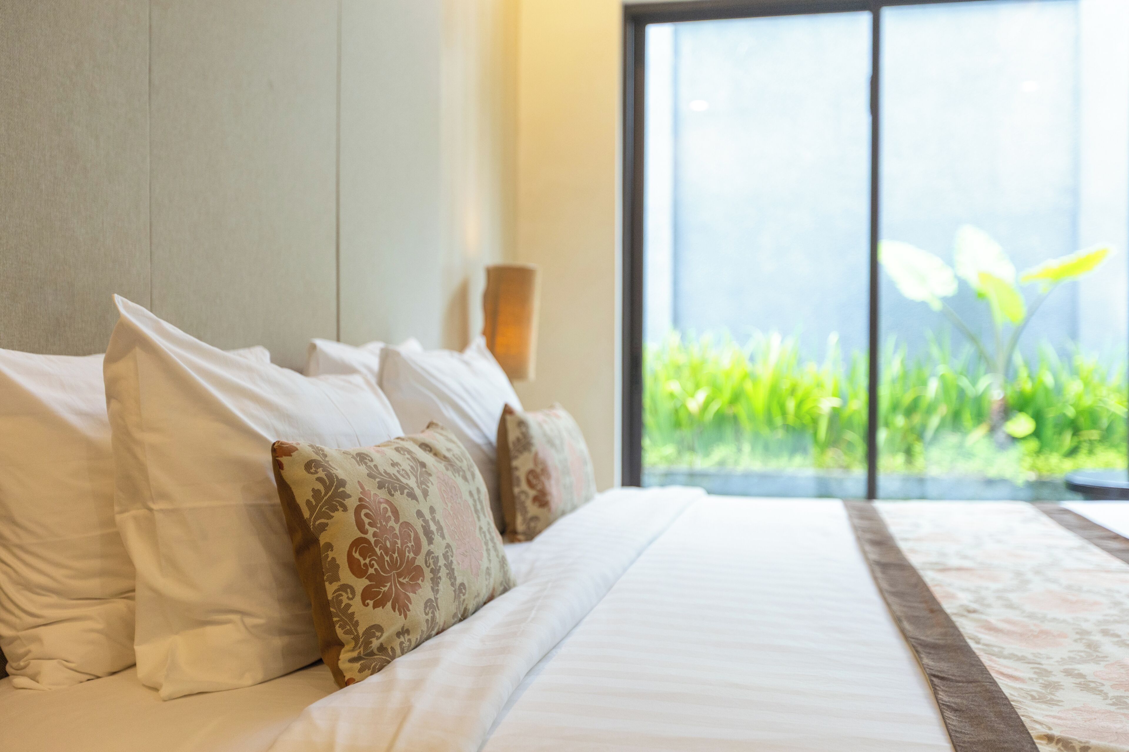 Una cama con almohadas blancas y estampadas, cabecera de madera y vistas a la vegetación a través de un ventanal amplio.