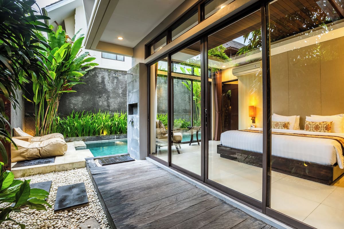 A modern bedroom with a large bed, a glass door leading to a pool, and a patio area with seating and plants.