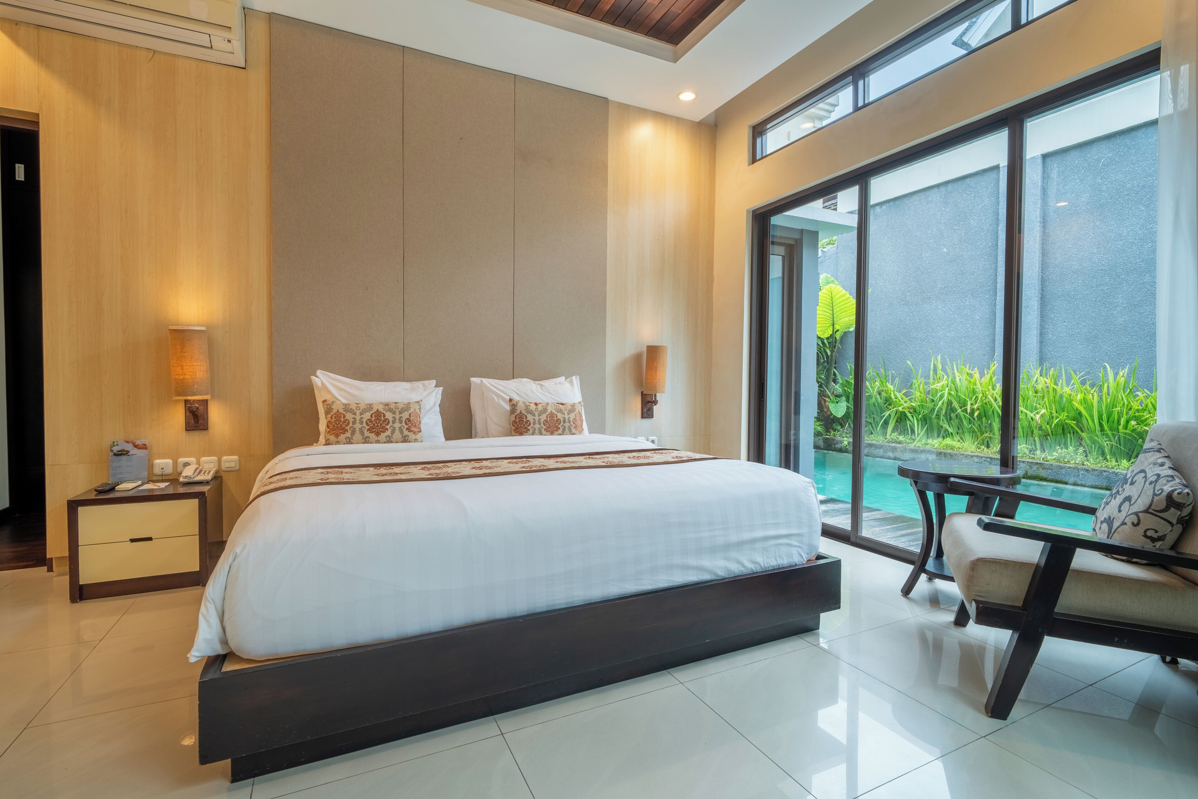 A modern bedroom with a large bed, bedside lamps, a nightstand, a view of a pool, and a seating area.