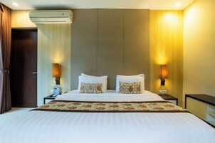 A hotel room with a large bed, two bedside lamps, an air conditioning unit, and a nightstand with a phone and a lamp.