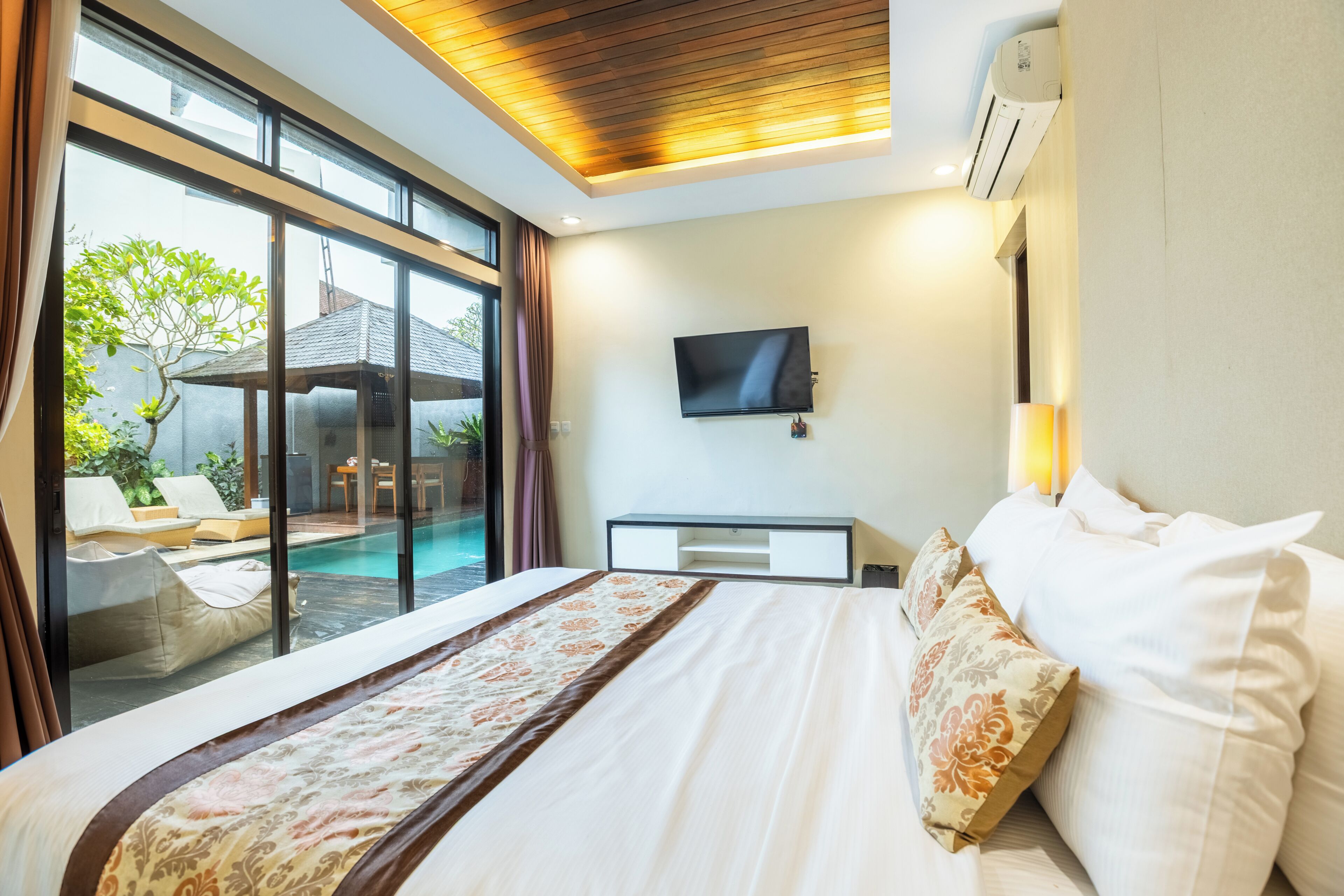 A hotel room with a large bed, a TV, a bedside table, and a view of a pool and outdoor seating area.