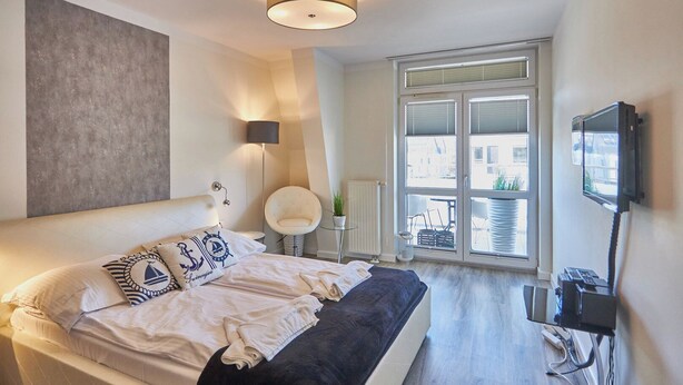 A modern bedroom with a large bed, a TV mounted on the wall, and a window with blinds. - Villa Mistral by Baltic Home (Swinoujscie)