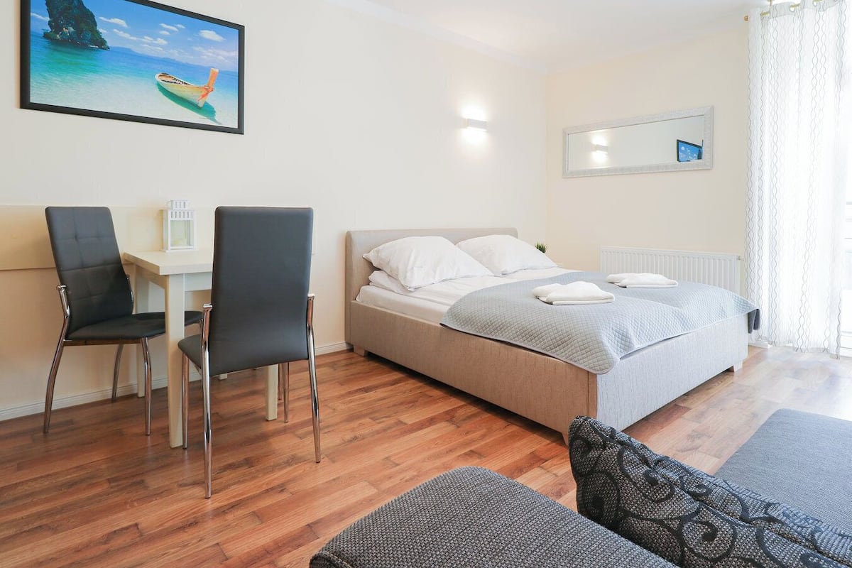A bedroom with a bed, two chairs, a desk, a window with curtains, and a painting of a boat.
