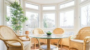 A corner room with a glass table surrounded by wicker chairs, a large plant, and a view of trees and power lines through the windows. - Villa Mistral by Baltic Home (Swinoujscie)