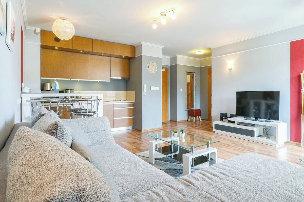 A modern living room with a sofa, a glass coffee table, and a TV. There is a kitchen area with wooden cabinets and a dining table.
