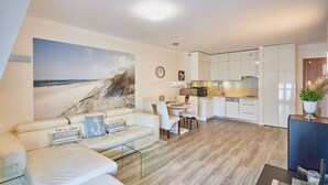 A modern living room with a large beach mural, a sofa with cushions, a glass coffee table, and a dining area with a kitchen in the background. - Villa Mistral by Baltic Home (Swinoujscie)