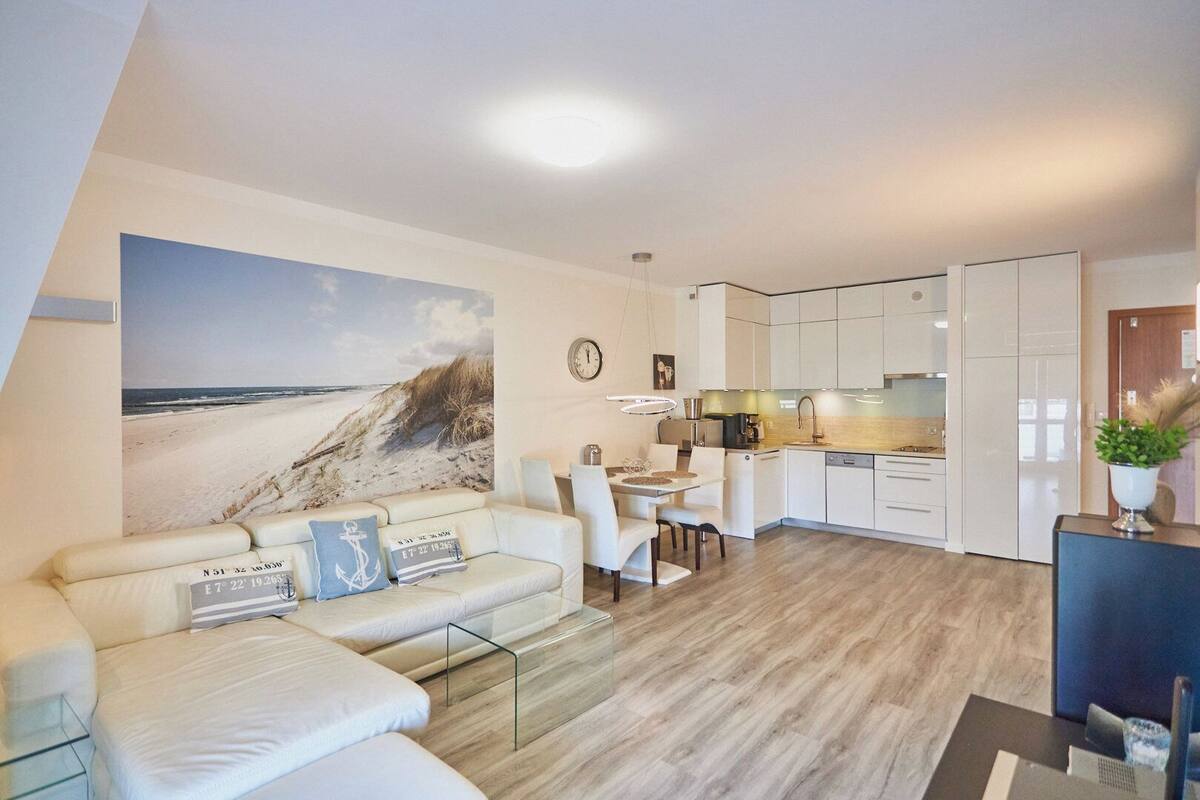 A modern living room with a large beach mural, a sofa with cushions, a glass coffee table, and a dining area with a kitchen in the background.