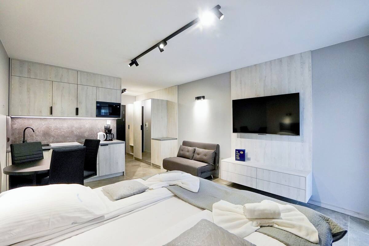 A modern hotel room with a bed, a desk, a chair, a sofa, a TV, and a kitchenette.