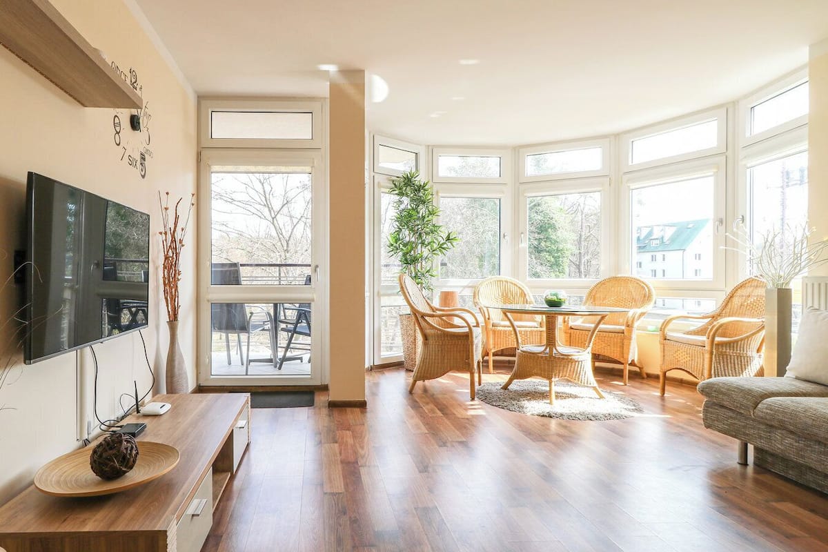 A spacious living room with a large window, a dining area with wicker chairs, a sofa, and a flat-screen TV mounted on the wall.