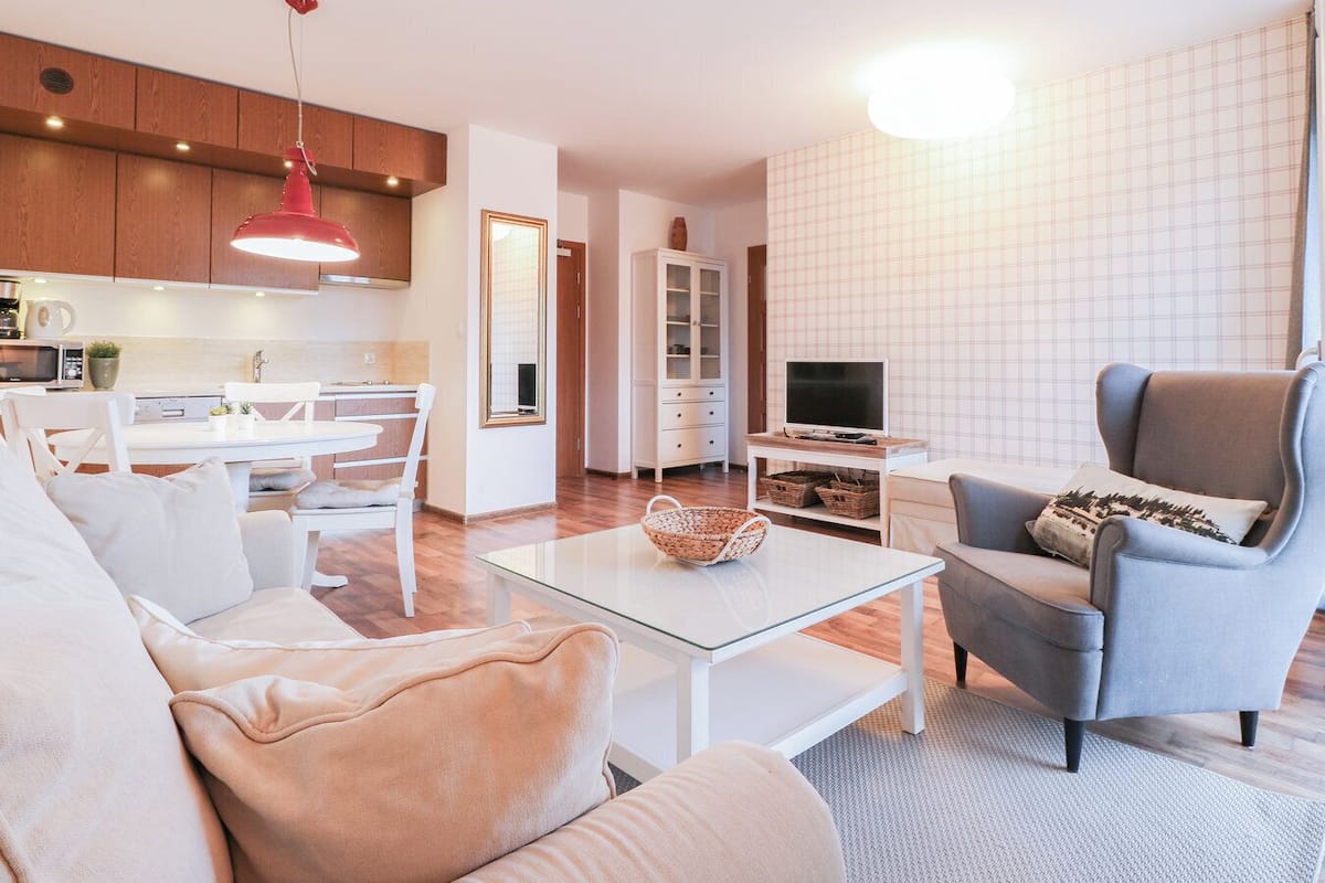 A modern living room with a sofa, armchair, and glass coffee table. A kitchen with wooden cabinets and a red pendant light is visible in the background.
