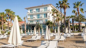 A beach with white lounge chairs and umbrellas, palm trees, and a hotel building in the background. - Hotel Caravelle Thalasso & Wellness (Diano Marina)