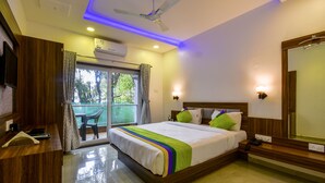 A modern hotel room with a large bed, a ceiling fan, an air conditioning unit, a balcony with a chair, and a wooden desk with a telephone. - Treebo Prince Palace With Swimming Pool (Mahabaleshwar)