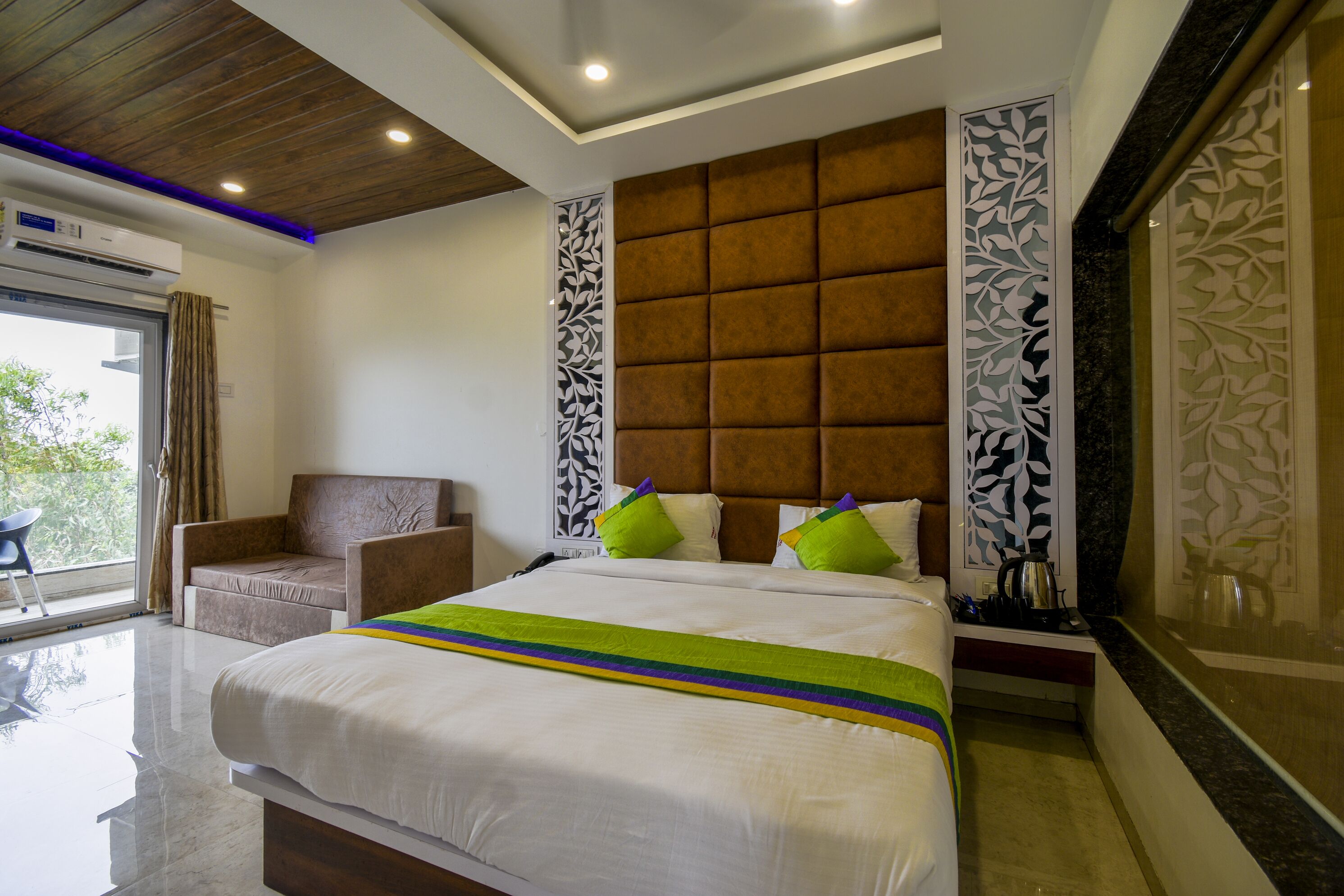 A modern hotel room with a large bed, a sofa, a decorative headboard, and a view of the outdoors.