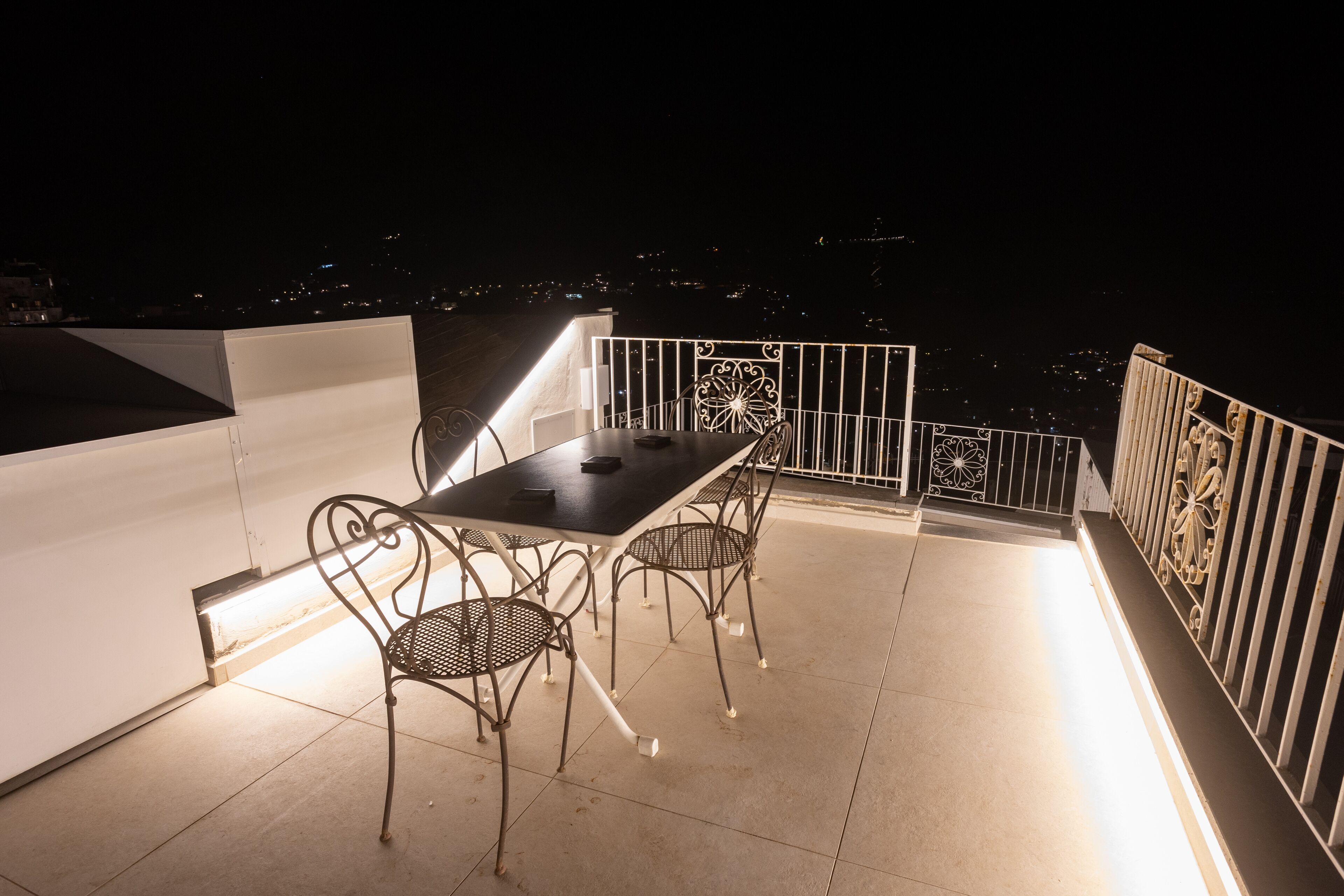 A rooftop terrace with a table and chairs, a railing, and a city view at night.