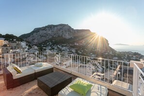 Un balcon doté de meubles en osier et offrant une vue sur une ville côtière et une chaîne de montagnes.