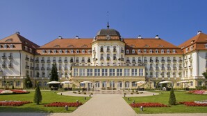 Um hotel grande e histórico com uma cúpula central, arquitetura simétrica e um jardim bem cuidado com caminhos e canteiros de flores.