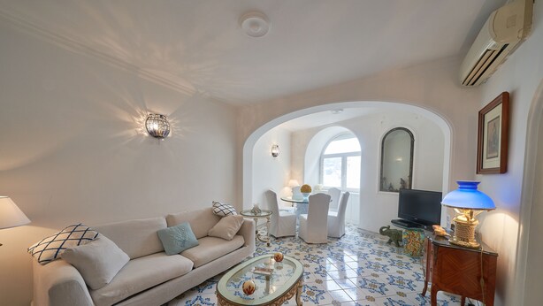 Una sala de estar con un sofá, una mesa de centro y un televisor. Hay un comedor con sillas y una mesa. El piso tiene baldosas con diseño.