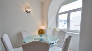 Un comedor con una mesa de vidrio, sillas blancas, un frutero, una lámpara de pared y una ventana con vistas a edificios.