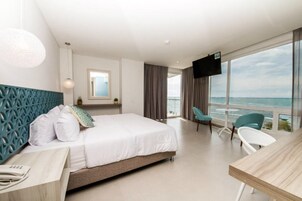 Habitación de hotel con cama, escritorio, silla, televisor y un ventanal con vista al océano.