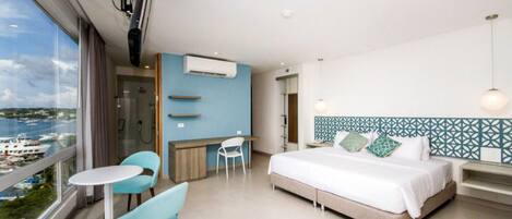 A modern hotel room with a large bed, a balcony with a view of the sea, a small desk, and a seating area.