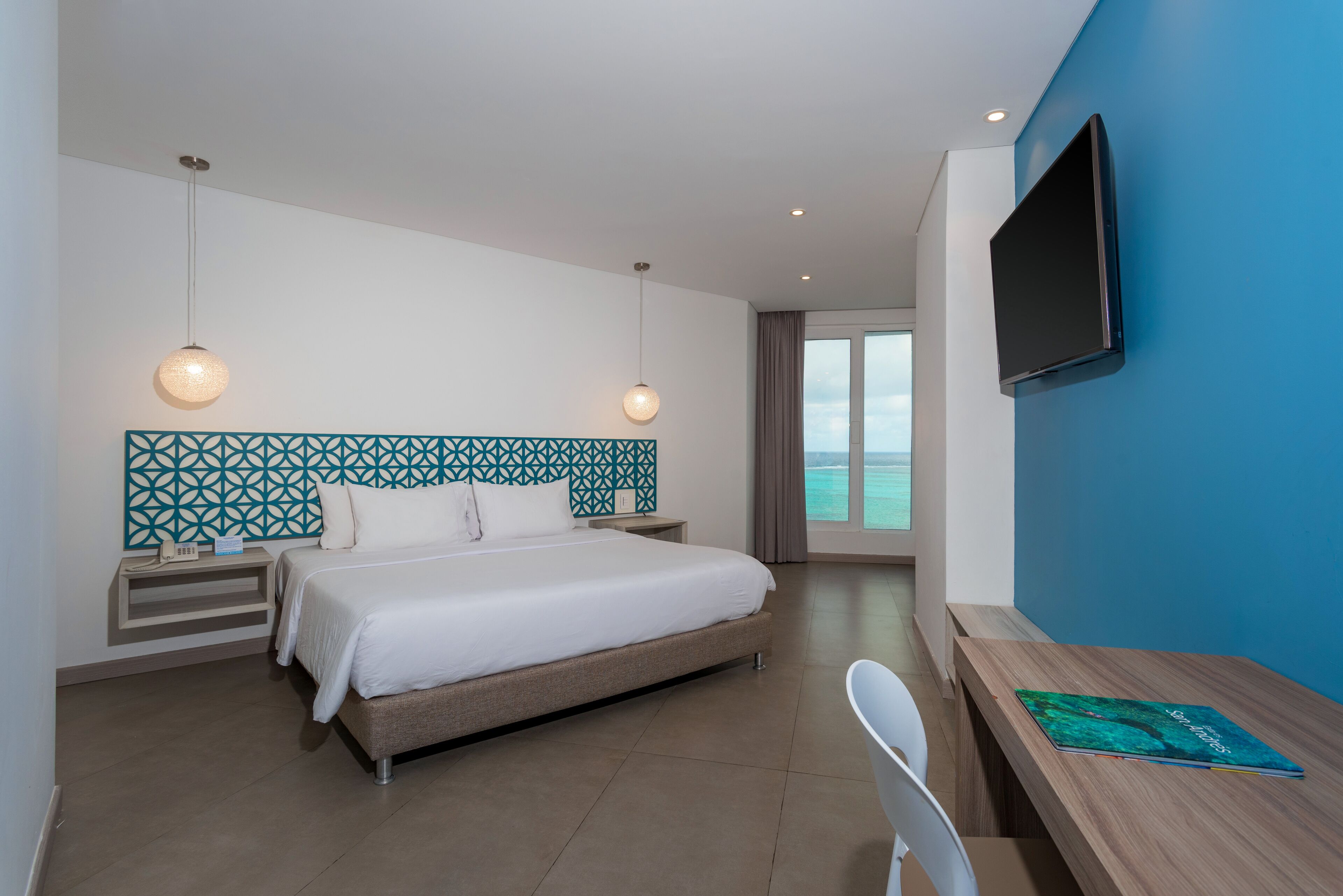 A modern hotel room with a large bed, a desk, a TV, and a view of the ocean.
