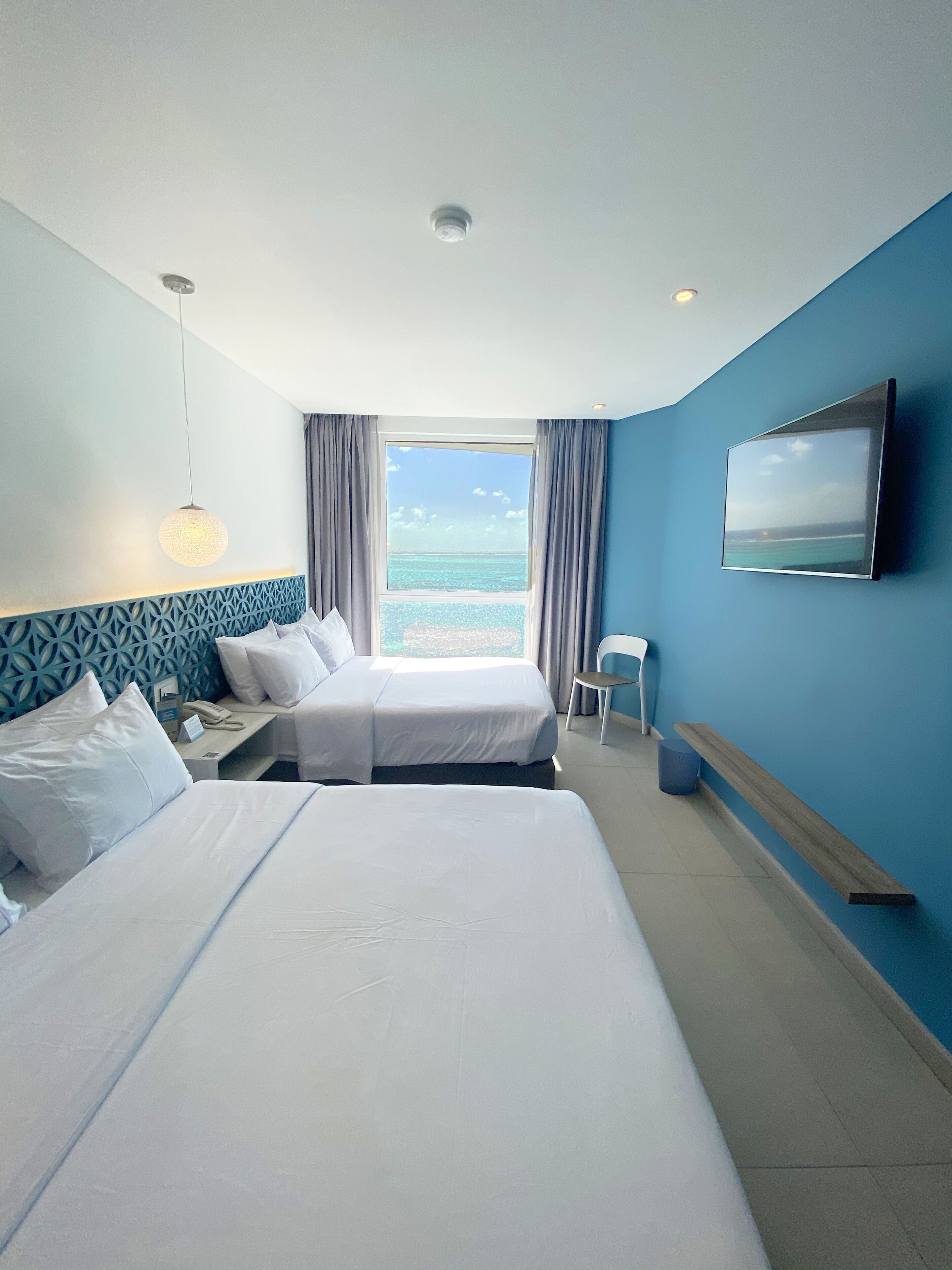 Une chambre d’hôtel avec deux lits, une grande fenêtre donnant sur la mer, un téléviseur fixé au mur et un luminaire au plafond.