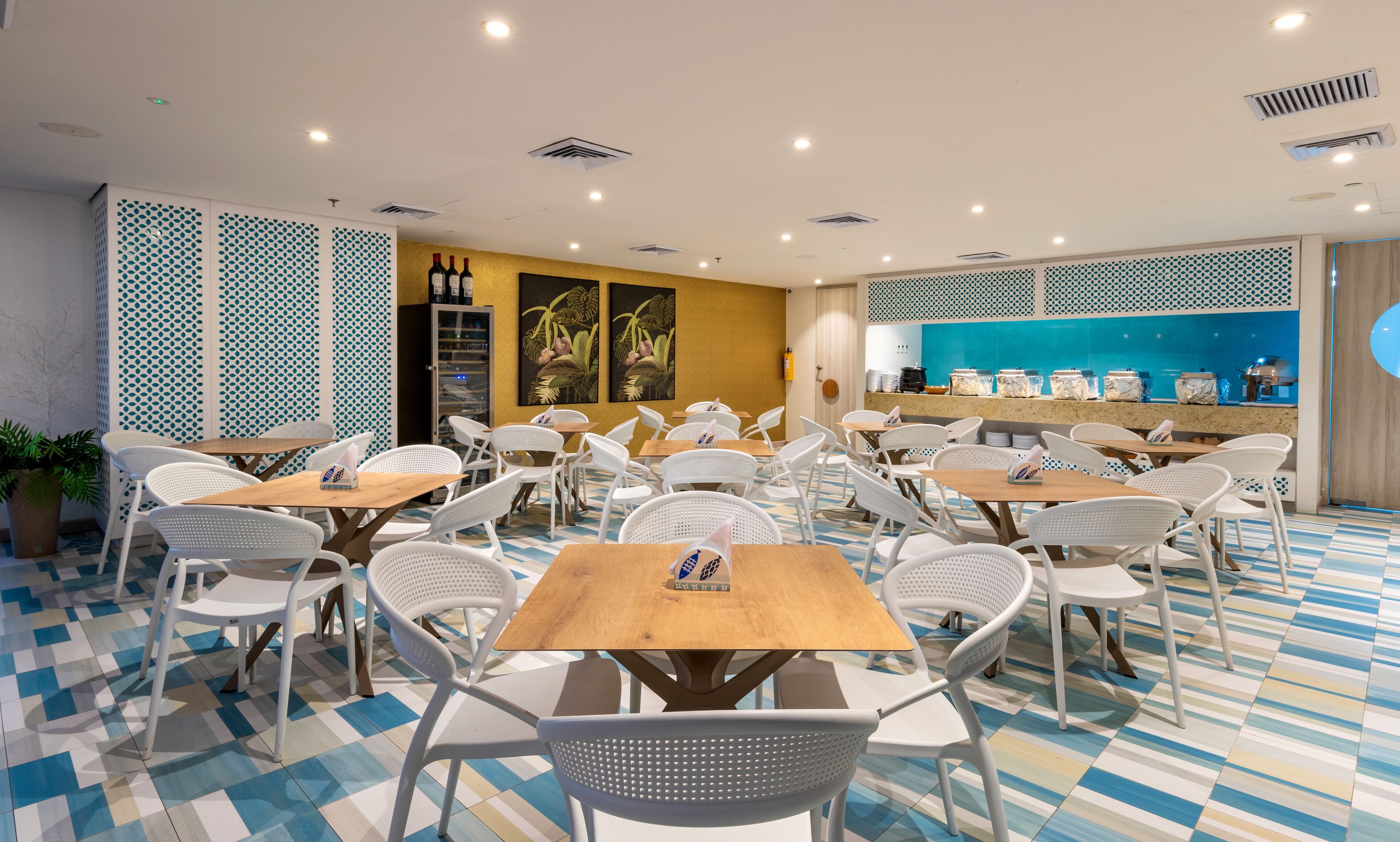 Un restaurante con sillas blancas y mesas de madera, suelo estampado y una zona de cocina con recipientes de comida.