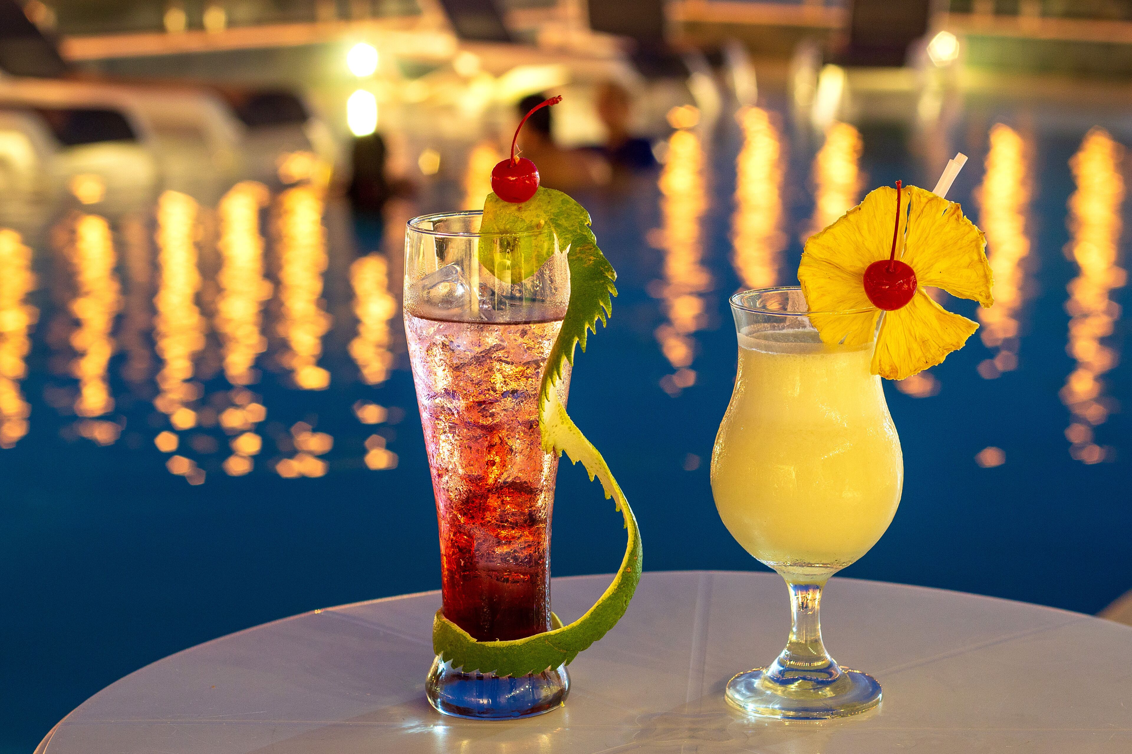 Two cocktails on a table by a pool with a blurred marina background.