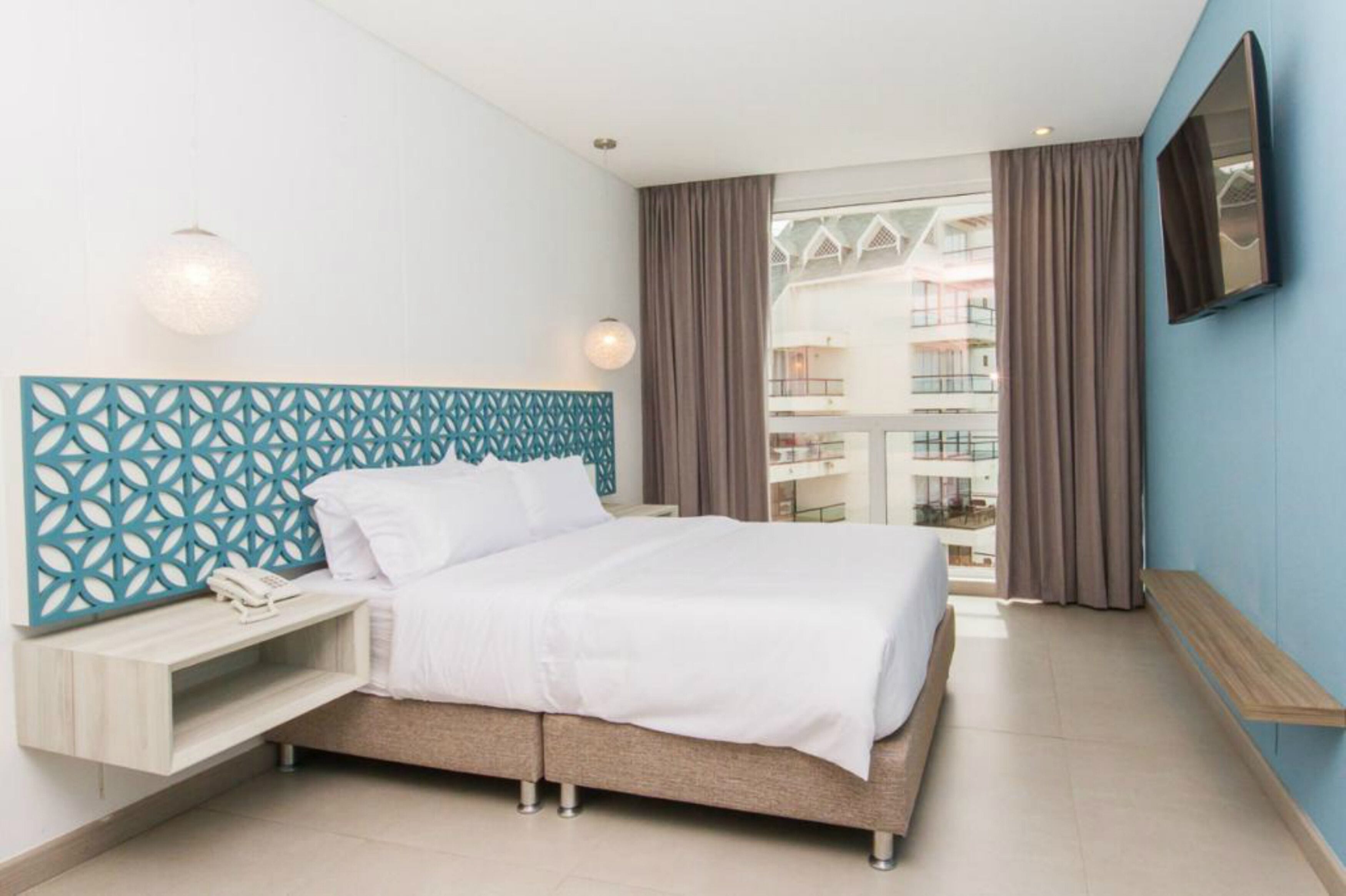 A modern hotel room with a bed, a nightstand, a phone, and a view of the building's exterior.