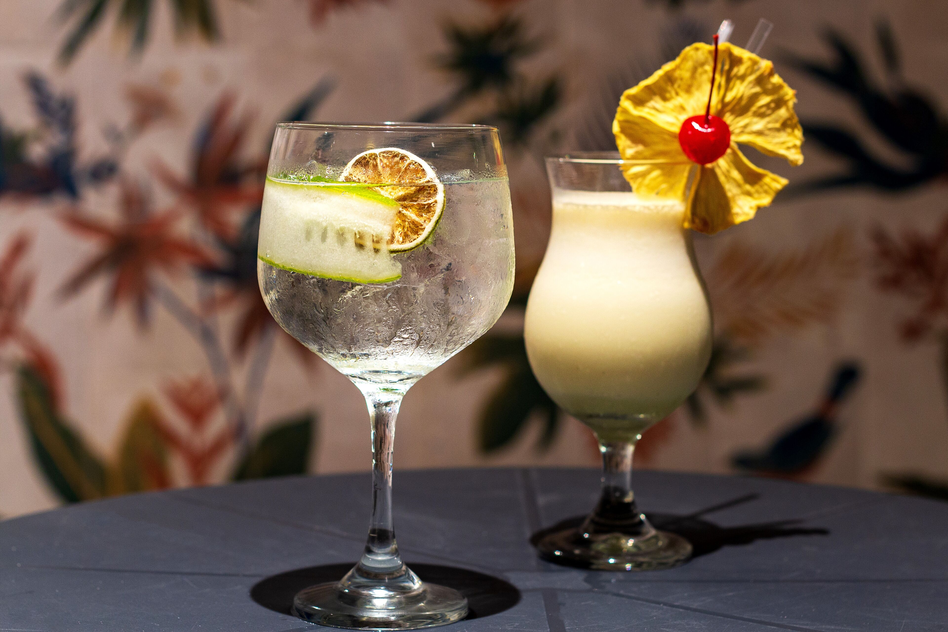 Dois cocktails sobre uma mesa, com papel de parede de tema tropical.