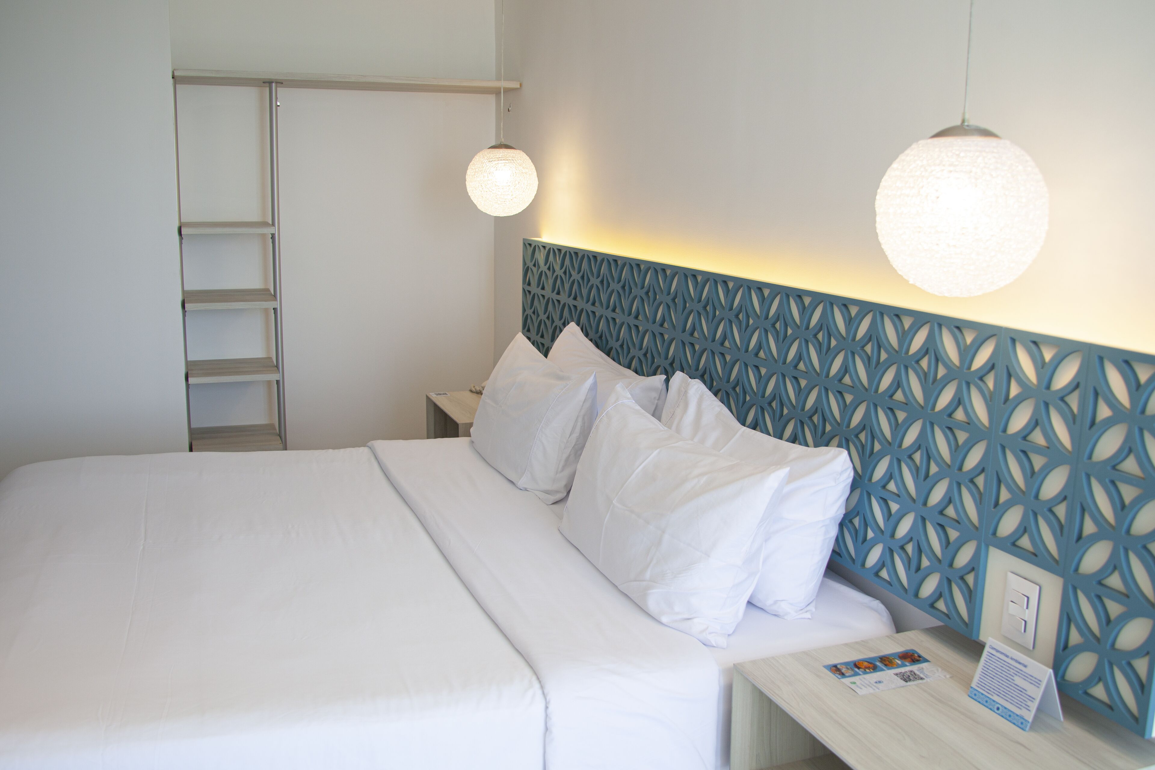 Una cama bien hecha con sábanas blancas, cabecera con estampado geométrico y mesita de noche con lámpara y folletos informativos.