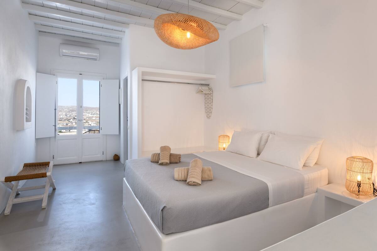 Une chambre à coucher au style minimaliste, avec un lit, des lampes de chevet et une vue sur l’extérieur, visible à travers une porte.
